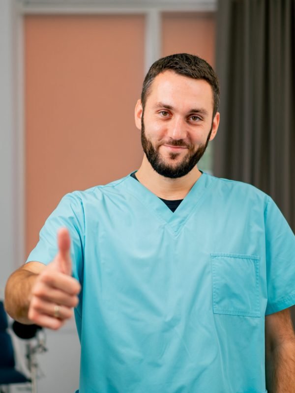 A portrait of a smiling male doctor with a beard standing in a medical office showing class sign and looking straight