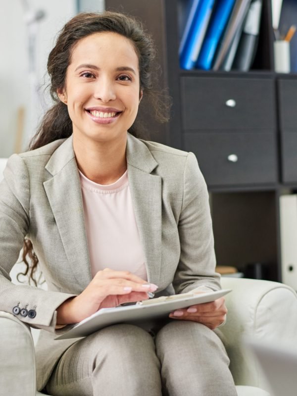 Positive successful beautiful mixed race psychologist with clipboard sitting in comfortable armchair and looking at camera in personal office