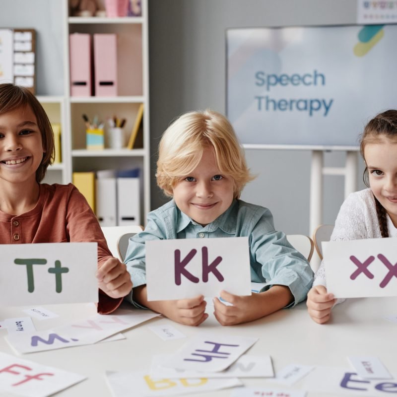 Portrait of group of children sitting at the table holding cards with English letters and smiling at camera