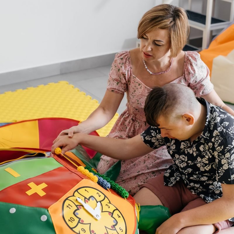 Children with disability getting sensory activity with toys, balls, small objects, cerebral palsy boy playing calming game, training fine motor skills. Rehabilitation center with therapist, mother