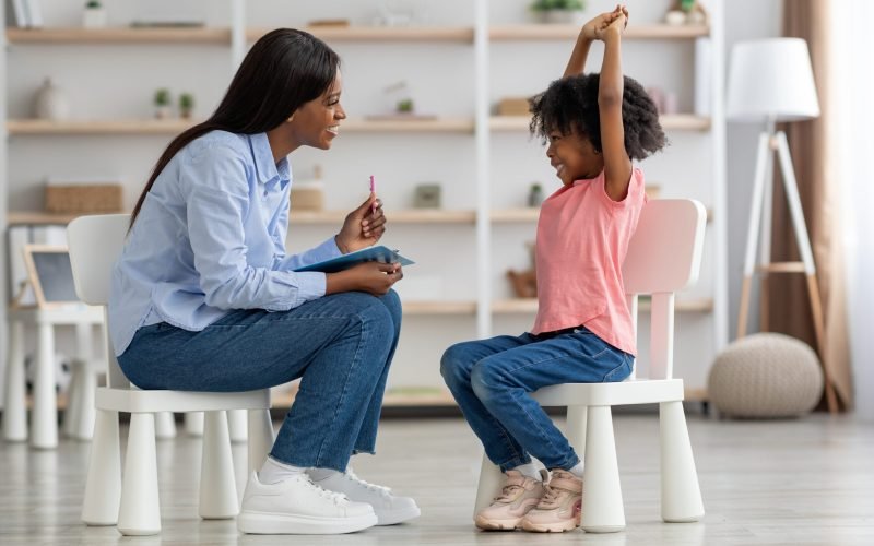 Excited cute little african american girl at child psychologists office, sitting on chair in front of attractive female therapist, raising hands up, smiling, feeling safe. Child psychotherapy concept