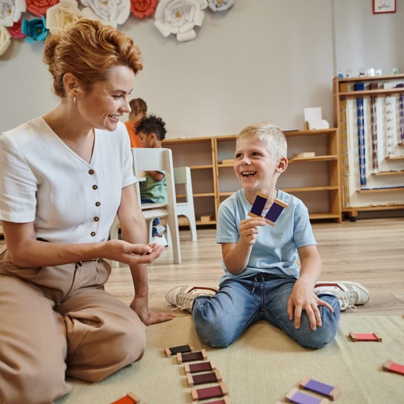 montessori school, happy boy playing color matching game near female teacher, sitting on floor